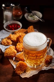 Beer with froth in glass mug near fried chicken nuggets served on creased paper on table with sauces and spices