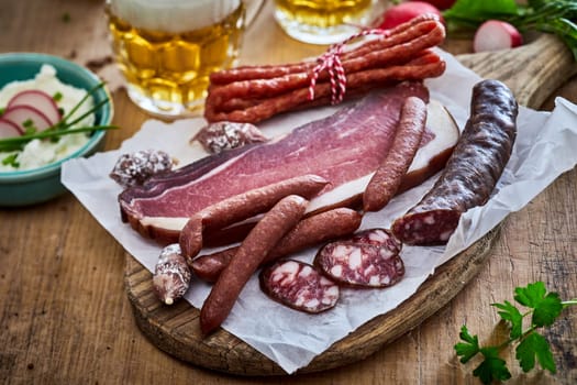 Smoked lard and assorted sausages served on wooden chopping board on table with hummus and mug of beer in kitchen