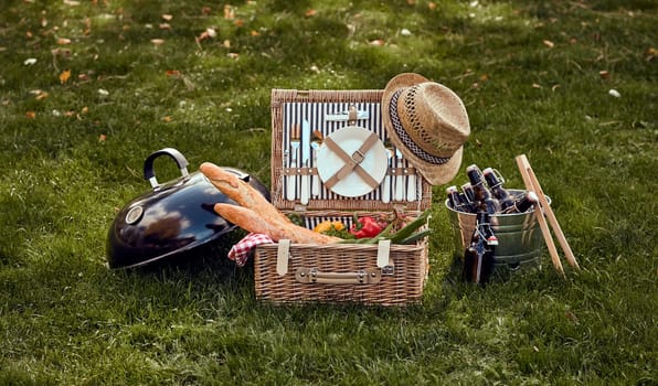 Wicker basket with tableware filled with baguettes and fresh vegetables placed on grassy lawn with bottles of alcohol drinks in bucket and grille lid during picnic