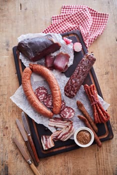 Top view of various cured sausages and smoked lard served on wooden table with knives and checkered napkin in kitchen