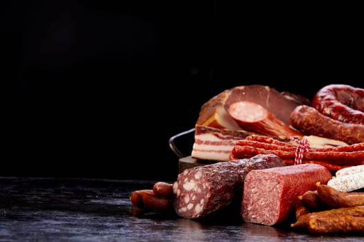 Assorted types of tasty cured sausages and delicious smoked lard placed on dark table in modern studio on black background