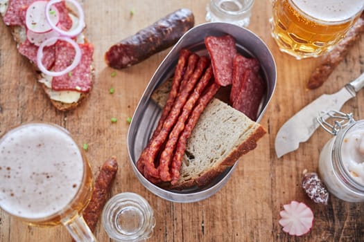 Top view of various types of sausages and bread served on wooden table with knife and mugs of beer in kitchen