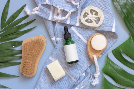 Green Dropper bottle, skin and hair care accessories on blue bath towel near tropical leaves top view, copy space. Packaging mockup. Natural homemade products