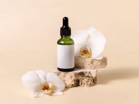 Green glass dropper bottle on stone near white orchid flowers on light yellow, close up, mockup. Skincare handmade beauty product, serum or lotion. Exotic natural cosmetics..
