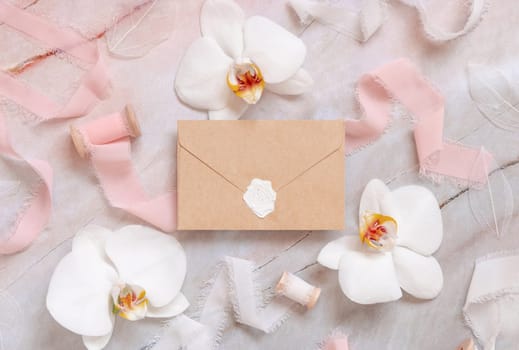 Wedding sealed envelope near white orchid flowers and silk ribbons on marble top view, mockup. Romantic scene flat lay. Valentines or Spring romantic concept