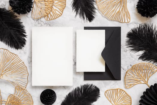 Blank cards and black envelope on marble table near black feathers and golden leaves top view. Wedding set mockup. Elegant femminine suite with paper cards, flat lay