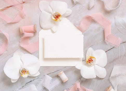 Wedding horizontal card and envelope near white orchid flowers and silk ribbons on marble top view, mockup. Romantic scene flat lay with handmade paper card