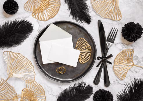 Black and golden wedding table setting with a place card and rings top view, mockup. Elegant flat lay with paper card and envelope, wedding or holiday stationery template