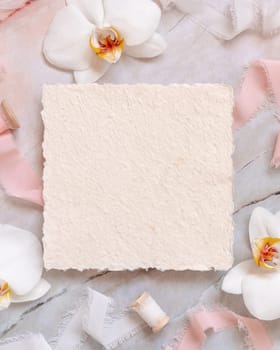 Wedding square card near white orchid flowers and silk ribbons on marble top view, mockup. Romantic scene flat lay with handmade paper card. Valentines or Spring romantic concept
