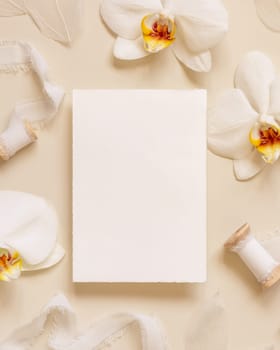 Wedding Card near white orchid flowers and silk ribbons on light yellow top view, mockup. Romantic scene with vertical blank card flat lay. Valentines or Spring romantic concept