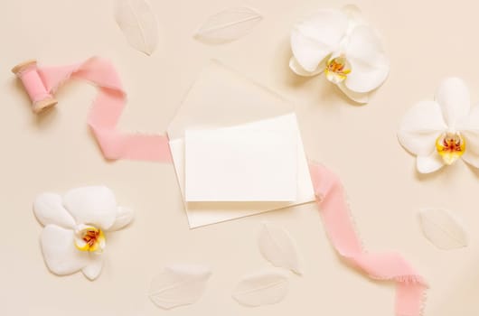 Wedding Card over envelope near white orchid flowers and silk ribbons on light yellow top view, mockup. Tropical scene with horizontal blank card flat lay