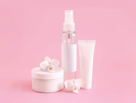 White cosmetic jar, tube and bottle near white orchid flower on light pink, close up, mockup. Skincare beauty product, cream or lotion. Exotic natural cosmetics, pastel minimal composition..