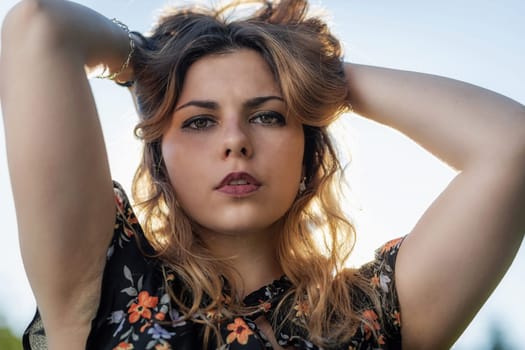 Atractive beautiful  young woman with both hands in her hair is posing outdoors against blue sky closeup.