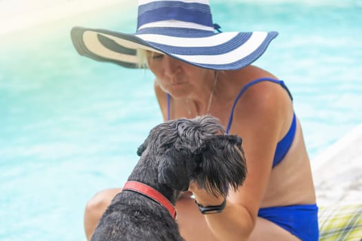 Woman wearing summer straw hat is posing with black schnauzer dog by the pool. Horizontally.