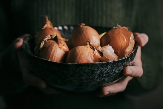 Crop unrecognizable person in casual clothes holding bowl with fresh ripe unpeeled onions