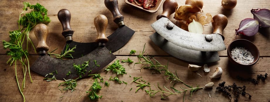 From above of traditional mezzaluna sharp knives placed on wooden table with scattered herbs during cooking process in light kitchen