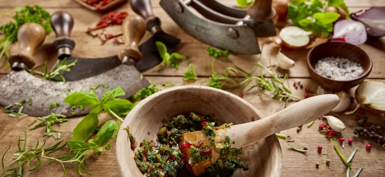 From above of wooden mortar with greens served on table with old fashioned chopping knives and scattered herbs in light kitchen