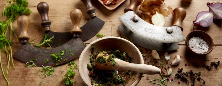 From above of mortar and pestle with grinded herbs placed on wooden table with old fashioned mezzaluna knives in kitchen