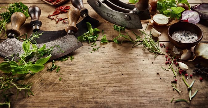 From above of traditional mezzaluna knives on wooden table with scattered fresh green herbs and various flavorings in light kitchen