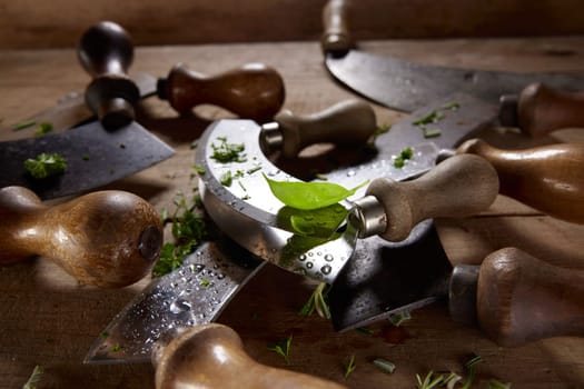 Green basil leaf placed on sharp metal blade of mezzaluna knife covered with freshly chopped herbs on wooden table in kitchen