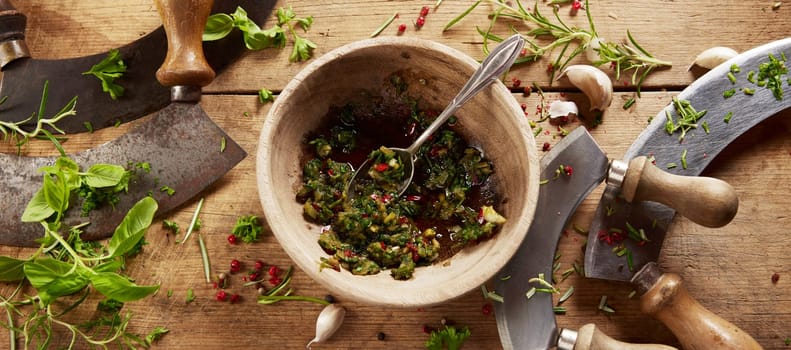 From above of mortar with freshly grinded herbs placed on wooden table with old fashioned traditional chopping knives in light kitchen