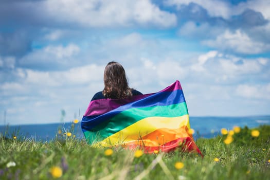 Woman sitting in a green meadow holding a Gay Rainbow Flag. Bisexual,gay, lesbian, transsexual symbol. Happiness, freedom and love concept for same sex couples