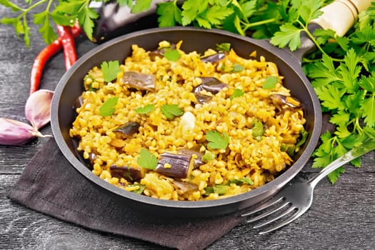 Bulgur stewed with eggplants, carrots, garlic and onions in a frying pan on a towel against the background of dark wooden board