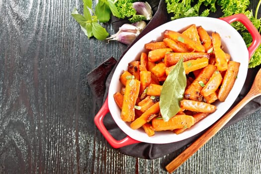 Carrots fried with garlic, bay leaf, spices and green onions in a pan on a napkin, basil and parsley on dark wooden board background from above