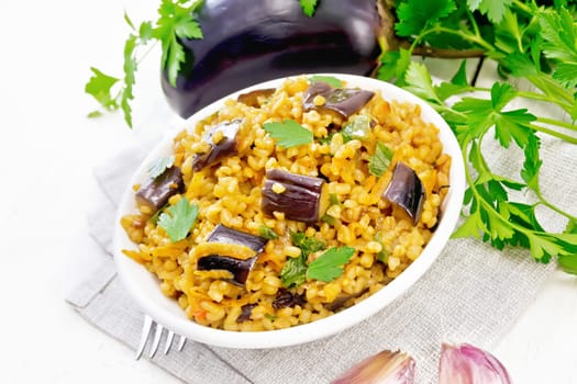 Bulgur stewed with eggplants, carrots, garlic and onions in a bowl on a towel against the background of light wooden board