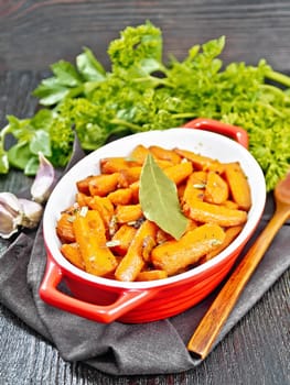 Carrots fried with garlic, bay leaf, spices and green onions in a pan on a towel, basil and parsley on dark wooden board background