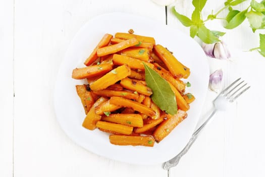 Carrots fried with garlic, bay leaf, spices and green onions in a plate, basil and napkin on light wooden board background from above
