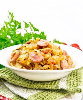 Cabbage stew with sausages in a white plate on a towel, tomatoes, parsley and a fork on the background of light wooden boards