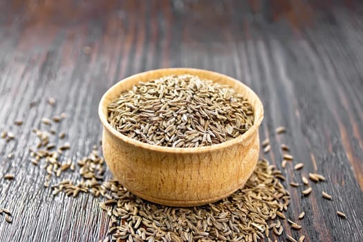 Cumin seeds in a bowl and on a table against dark wooden board background