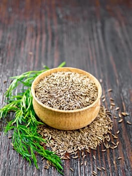Cumin seeds in a bowl and on table, a green twig of plant on background of wooden board