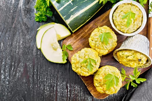 Small cheese and zucchini muffins with herbs, parsley, thyme, knife and towel on a dark wooden board background from above