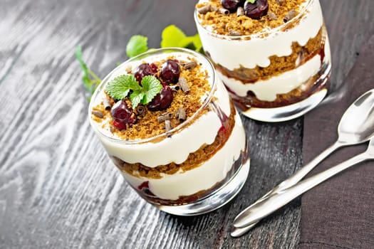 Dessert Black Forest of cherries, chocolate biscuit and soft cottage cheese with cream in two glasses, a towel, mint and a spoon on black wooden board background