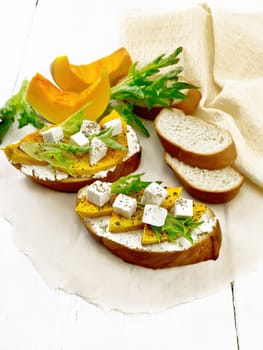 Bruschetta with baked pumpkin, salted feta cheese, ricotta, arugula and spices on parchment, towel and vegetable slices on the background of light wooden board