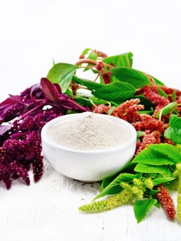 Amaranth flour in a bowl, brown, green and purple flowers of a plant on light wooden board background