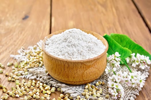 Buckwheat flour from green cereals in a bowl on sacking, groats on the table, flowers and buckwheat leaves on the background of a wooden board