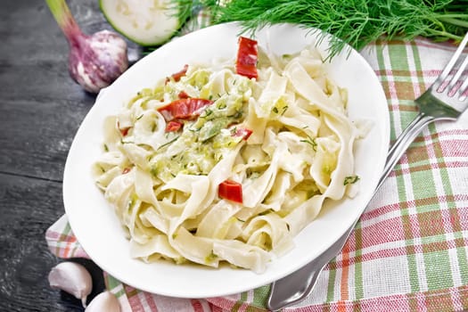 Fettuccine pasta with zucchini and hot red pepper in creamy sauce in white plate on a towel, garlic and a fork on wooden board background