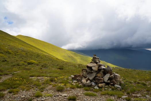 closeup of pyramid of stacked stones used for hiking trail tourist guidance with beautiful sunny mountain slope range covered in green grass in the background and cloudy sky