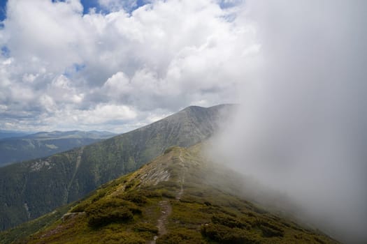landscape with green mountain peak with cloud mist passing by and trail going on down the ridge