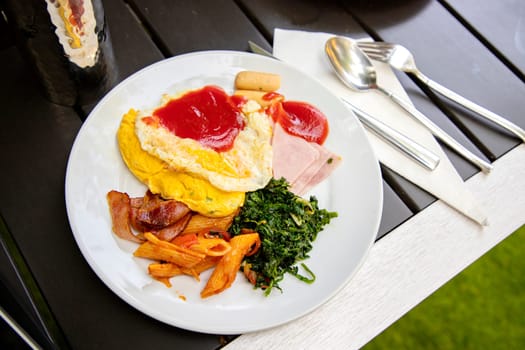 Breakfast from the buffet in a round white plate. on a modern wooden table There is a glass of water upside down, reflecting on it. There are stainless steel knives, spoons, forks placed beside them.