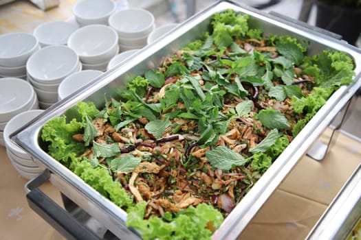 Larb Moo, sprinkled on top with mint leaves in buffet line. The container is a rectangular stainless steel tray with small white cups arranged on the side. Selective focus.