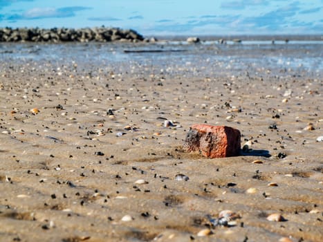 Low-angle landscape view of the German North Sea beach at low tide with a brick fragment in the foreground, a stone groyne in the blurred middle ground and a view to the horizon.