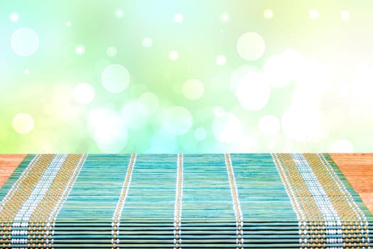 A green yellow bamboo mat on a rustic wooden table over bright blurred abstract background. Template for your product display montage. Space for design.