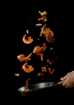 Crop anonymous male chef tossing food in frying pan while preparing delicious prawns and vegetables against black background