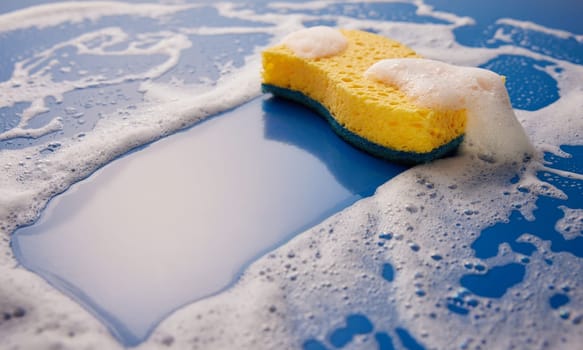 Yellow wash sponge for daily cleaning routine placed on foamy wet blue background covered with soap bubbles in light room