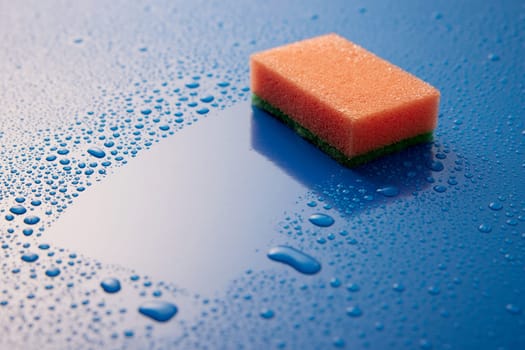 Orange scrub sponge placed on wet blue surface covered with small drops of water in light room during daily hygiene routine