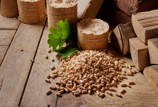 From above of pile of fuel pellets and fresh green oak leaves on wooden table with compressed biomass briquettes and woodpile